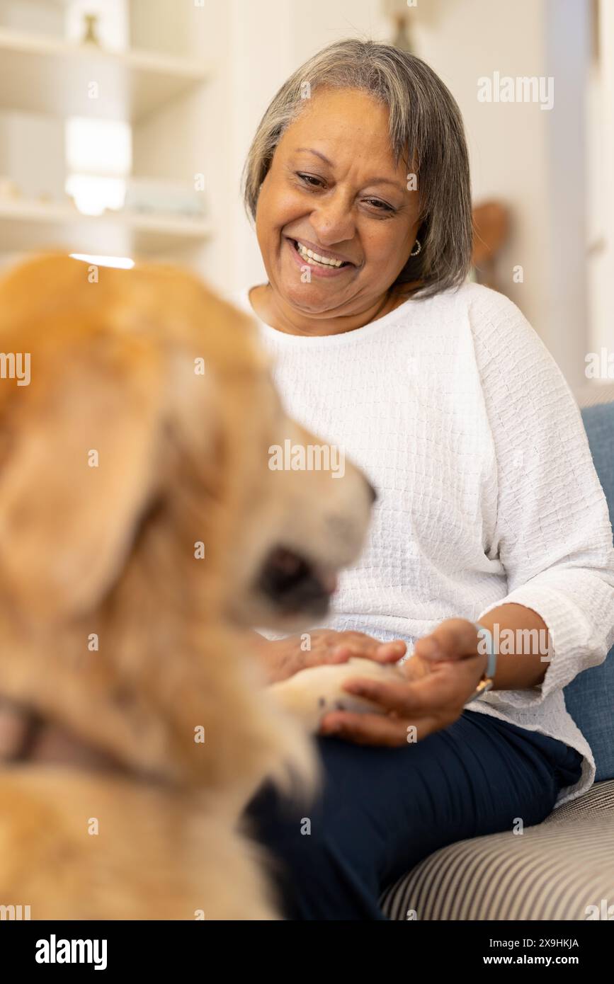 Zu Hause lächelt eine zweirassige Seniorin, während sie mit dem Hund auf einer Couch interagiert Stockfoto