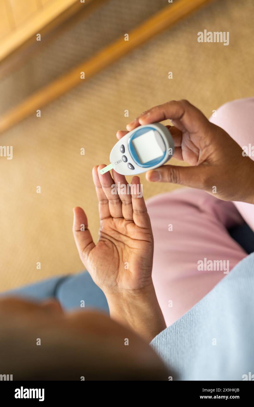 Zu Hause, zweirassige Seniorin, die den Blutzuckerspiegel mit Glukometer überprüft. Sitzen auf Couch in einem gemütlichen Wohnzimmer mit Teppichboden und Holzfu Stockfoto