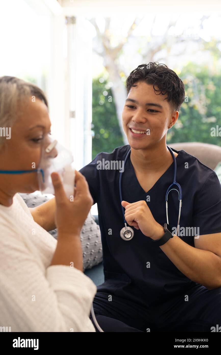 Zu Hause, junge birassische männliche Krankenschwester, die älteren weiblichen Patienten hilft. Sie sind in einem hellen Raum mit großen Fenstern, Grün draußen, was eine ruhige Atmosphäre schafft Stockfoto
