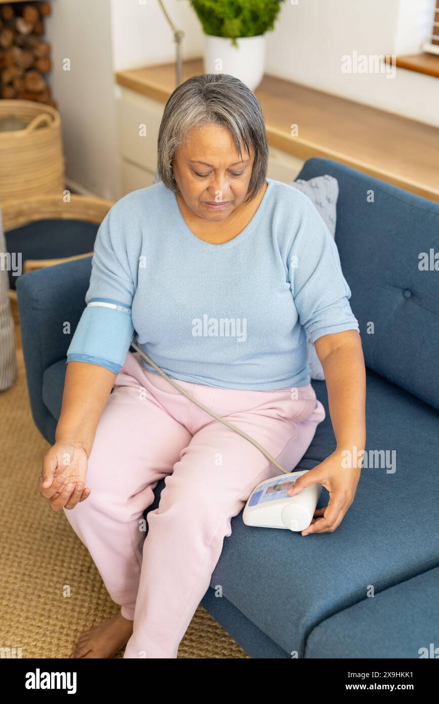 Zu Hause überprüft eine zweirassige Seniorin den Blutdruck mit einem Monitor am Arm. Sitzen Sie auf einem blauen Sofa in einem gemütlichen, gut beleuchteten Wohnzimmer mit einem gewebten ru Stockfoto