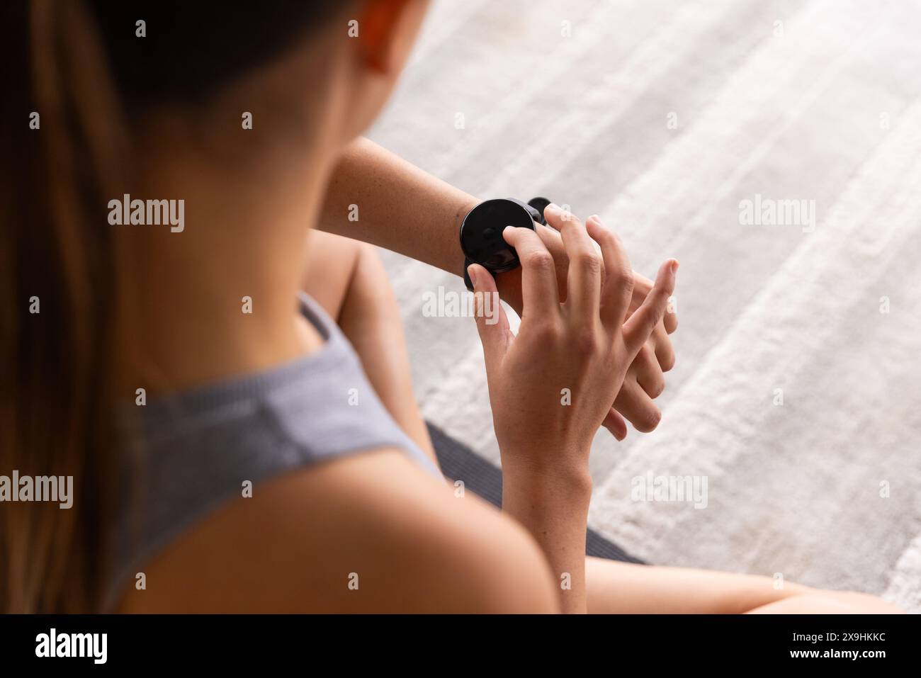 Zu Hause, junge kaukasische Frau, die Smartwatch benutzt, während sie auf einer Yogamatte sitzt, Kopierraum. Gemütliches Zuhause mit einem weichen, hellen Teppich, der noch dazu beiträgt Stockfoto