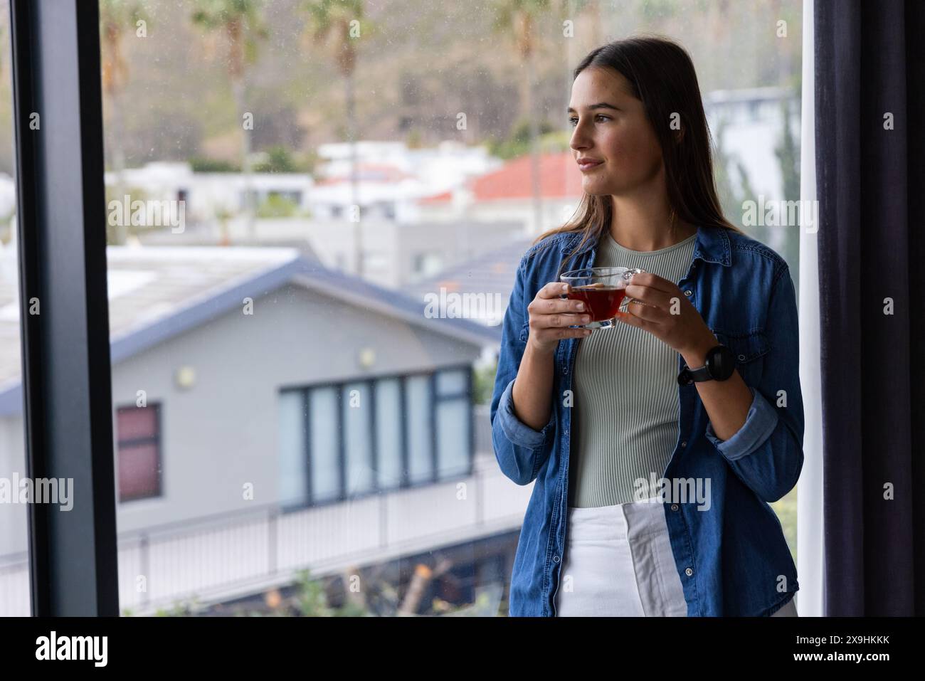 Zu Hause genießt eine junge kaukasische Frau eine Tasse Tee, während sie aus dem Fenster blickt. Sie ist in einem gemütlichen Heim-Spa mit einem ruhigen Blick auf die Nachbarschaft Stockfoto