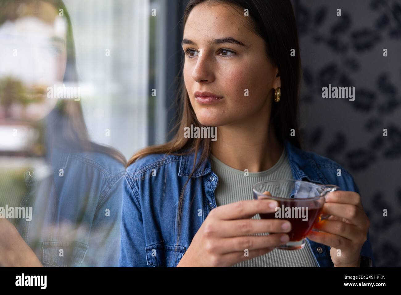 Zu Hause, eine junge kaukasische Frau, die eine Tasse Tee hält und nachdenklich aus dem Fenster blickt. Sie befindet sich in einer gemütlichen Spa-Umgebung mit sanfter Beleuchtung und Ruhe Stockfoto