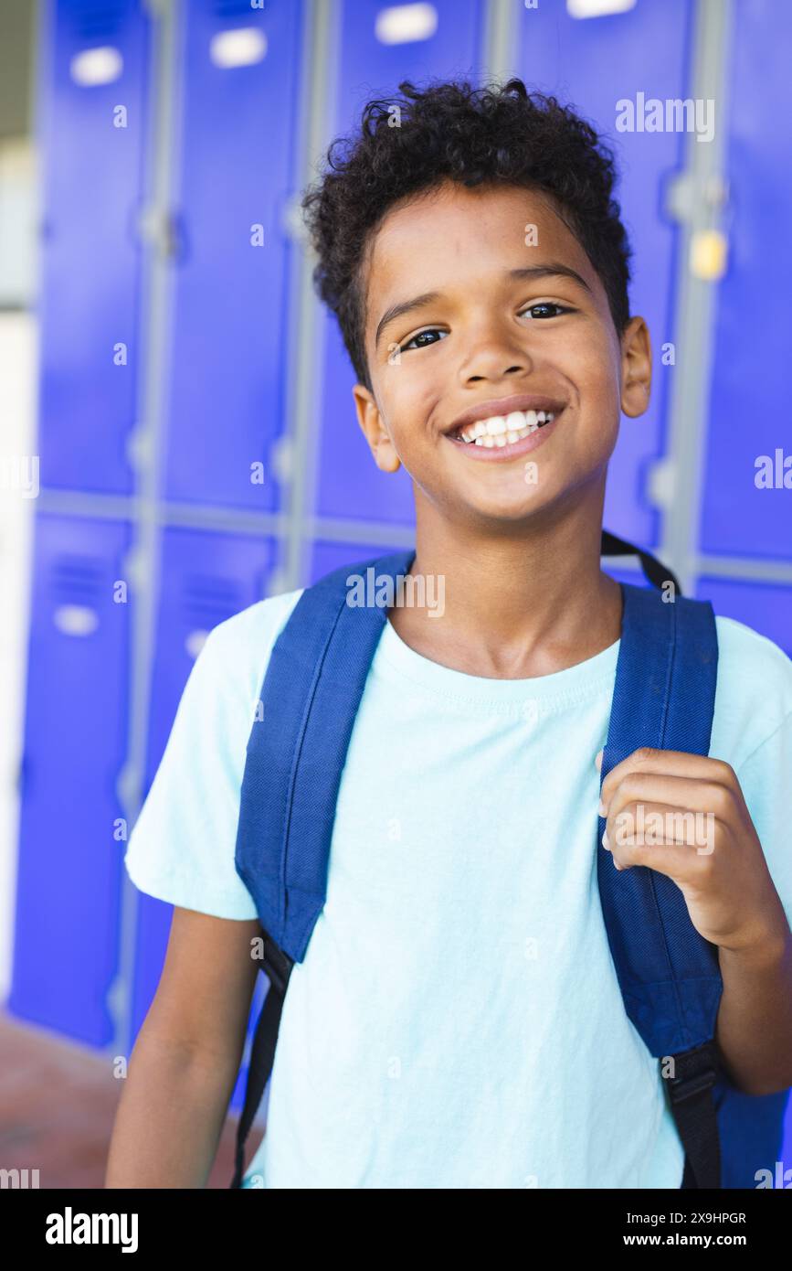Birassisches Kind mit einem hellen Lächeln steht vor blauen Schließfächern in der Schule. Mit hellblauem T-Shirt und Rucksack strahlt er Selbstvertrauen aus Stockfoto