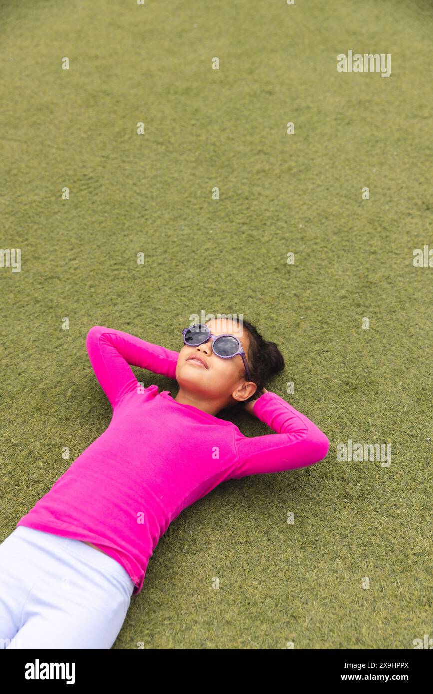 In der Schule, draußen, ein birassisches junges Mädchen mit Sonnenbrille, das auf Gras liegt und einen Kopierraum hat Stockfoto