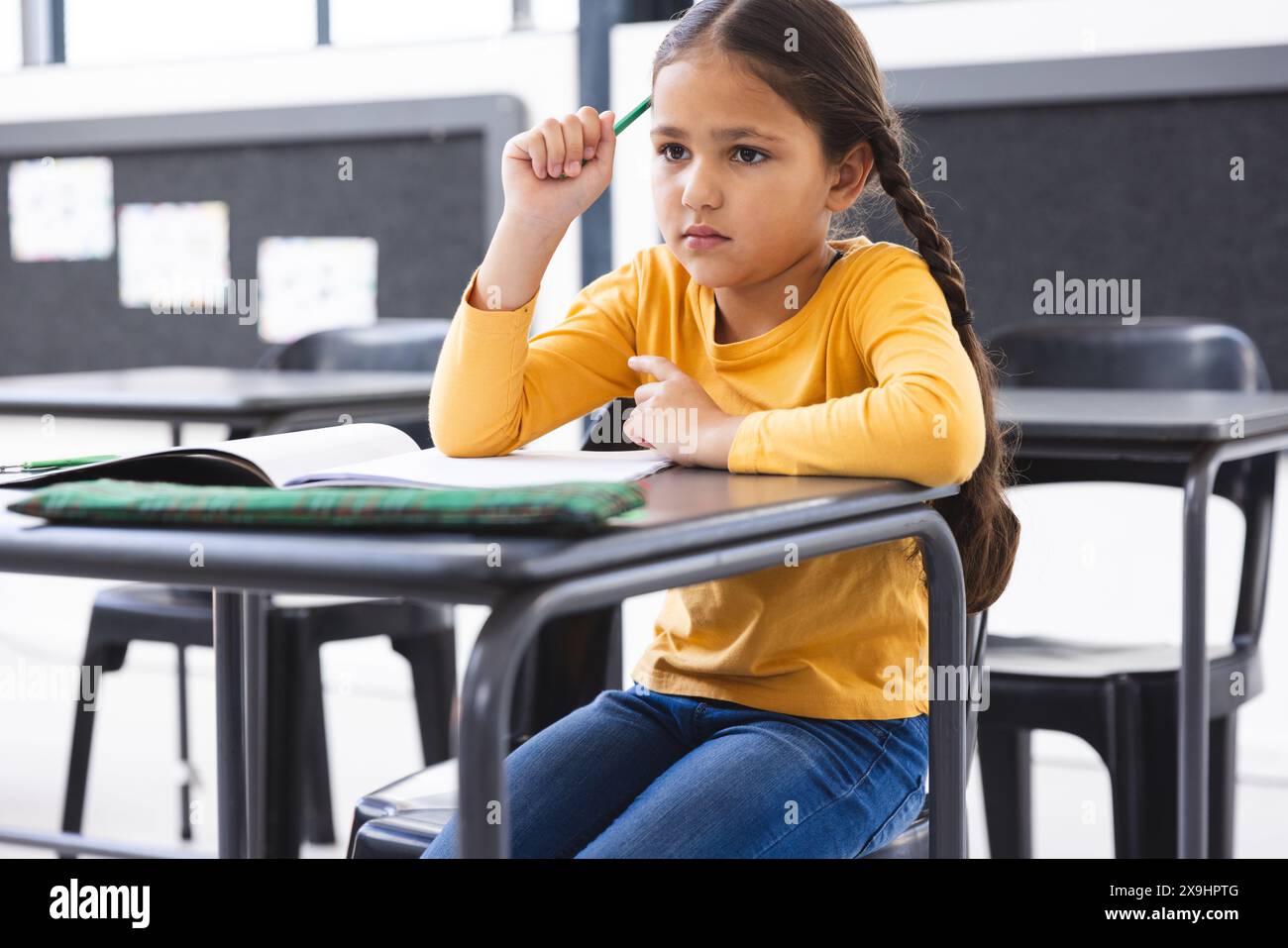 In der Schule sitzt ein junges birassisches Mädchen an einem Schreibtisch in einem Klassenzimmer und sieht nachdenklich aus Stockfoto
