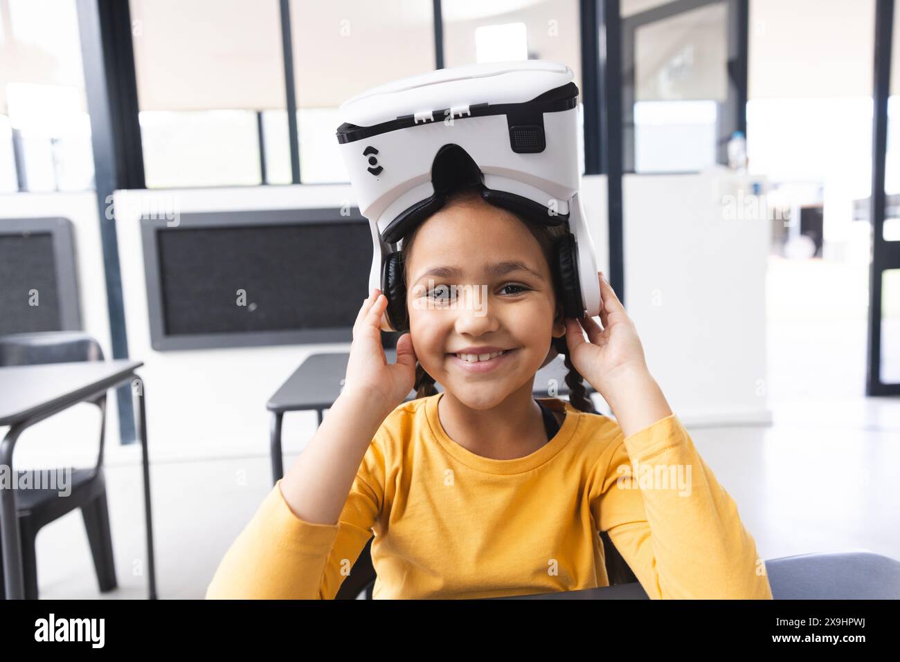 In der Schule, im Klassenzimmer, lächelt ein junges birassisches Mädchen, das ein VR-Headset trägt Stockfoto