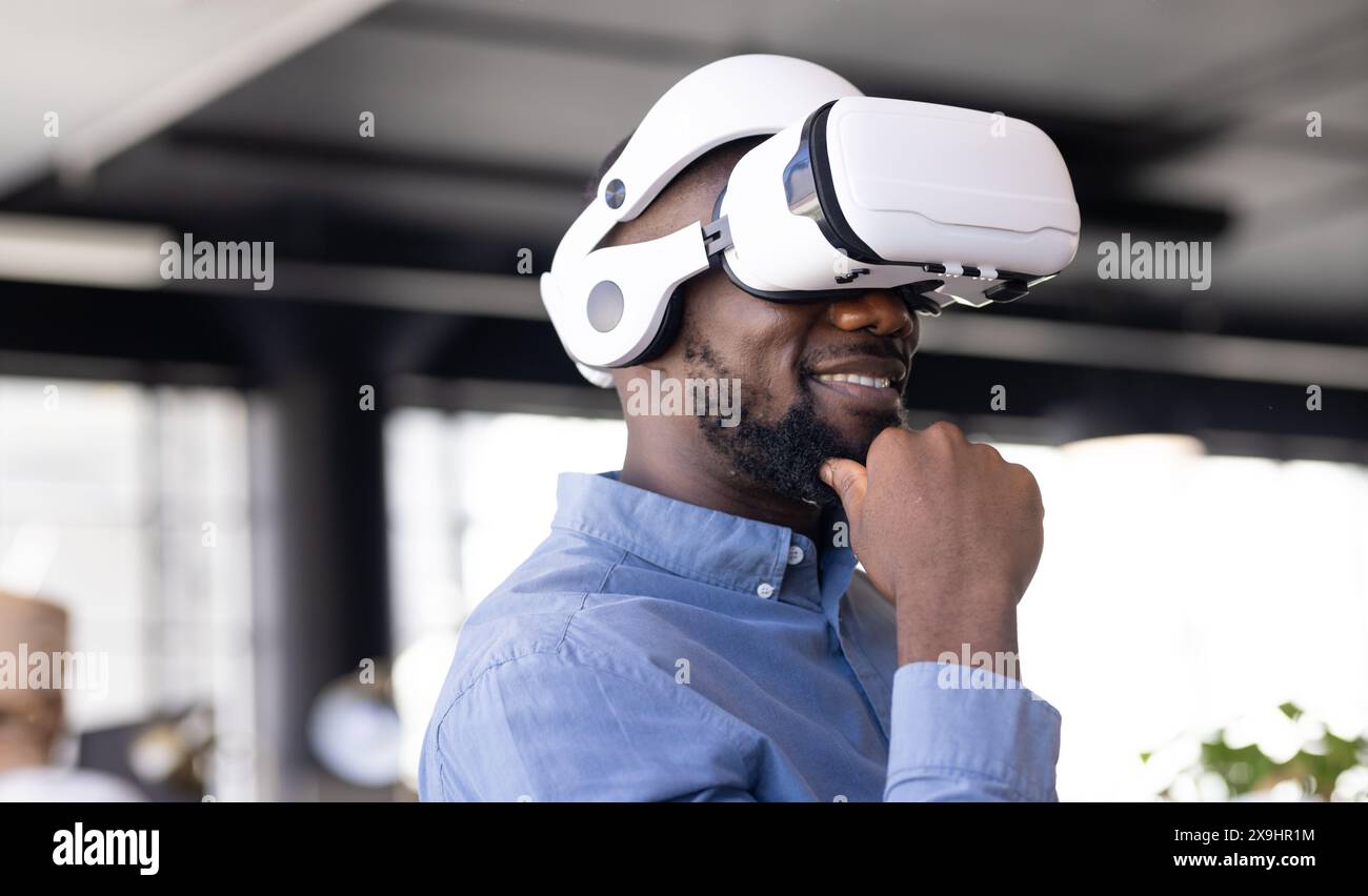 Junger afroamerikanischer Mann mit Virtual-Reality-Headset im Büro. Er hat kurze schwarze Haare, Bart und lächelt, während er sein Kinn berührt Stockfoto