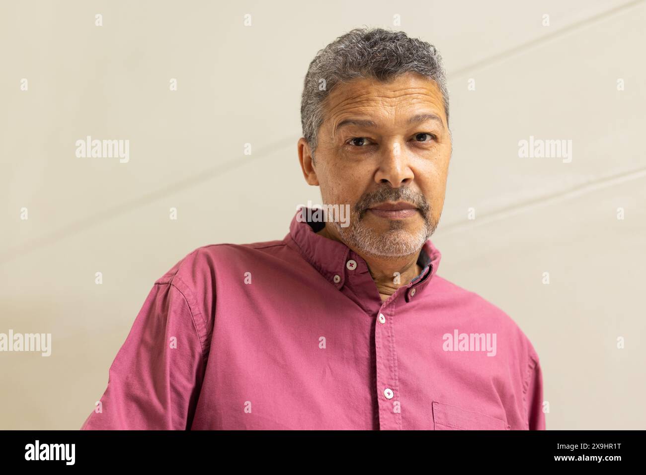 Zweirassiger älterer Mann mit kurzen grauen Haaren und beschnittenem Bart in einem rosa Hemd zu Hause, mit Blick in die Kamera. Er hat einen nachdenklichen Ausdruck, der wieder steht Stockfoto