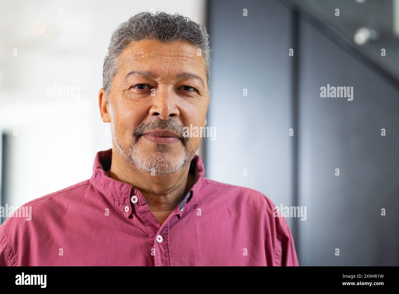 Zu Hause, zweirassiger älterer Mann mit kurzen, lockigen, grauen Haaren und Bart, der drinnen steht. Mit rosa Button-up-Hemd und einem ruhigen Blick in die Kamera Stockfoto