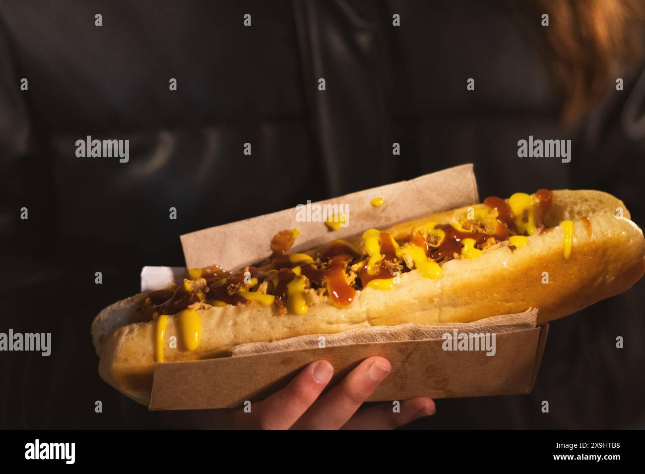 Anonyme Frau mit Streetstyle gefülltem Hotdog mit Belägen in der Hand in der Verpackung und in beleuchtetem Licht gegen dunkle Jacke Stockfoto