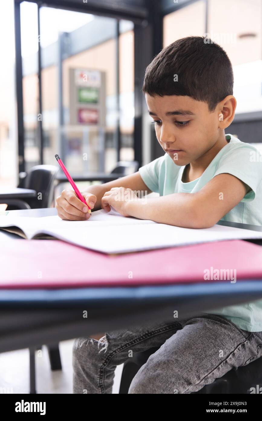 In der Schule, im Klassenzimmer, junger birassischer Junge, der sich darauf konzentriert, in einem Notizbuch zu schreiben Stockfoto