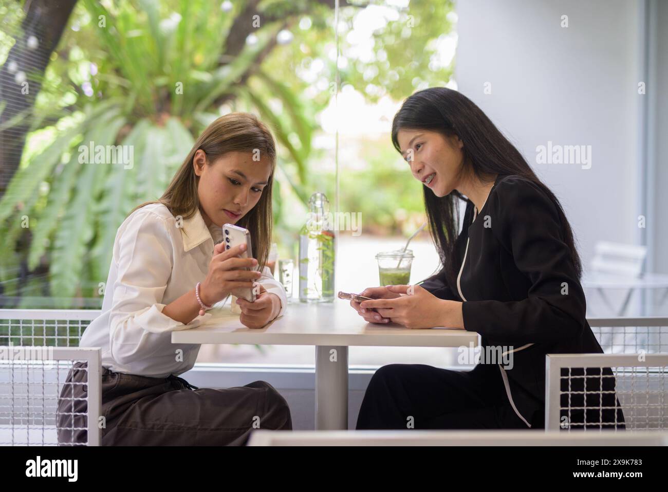 Zwei asiatische Frauen, die sich bei einem Kaffee in einem modernen Café mit viel Grün unterhalten. Stockfoto