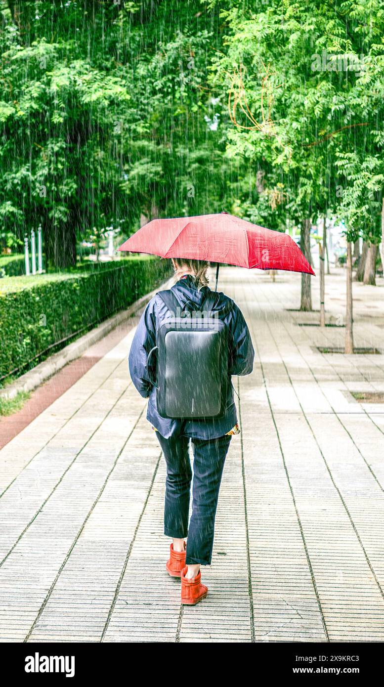 Eine Frau, die an einem regnerischen Tag unter einem roten Regenschirm läuft, einen schwarzen Rucksack und rote Schuhe trägt. Stockfoto