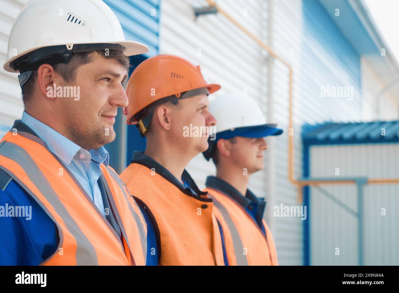 Drei Baumeister in Helmen und Westen stehen in Reihe. Freundliches Bauteam mit kaukasischem Aussehen. Porträts junger Arbeiter auf der Baustelle am Sommertag. Stockfoto