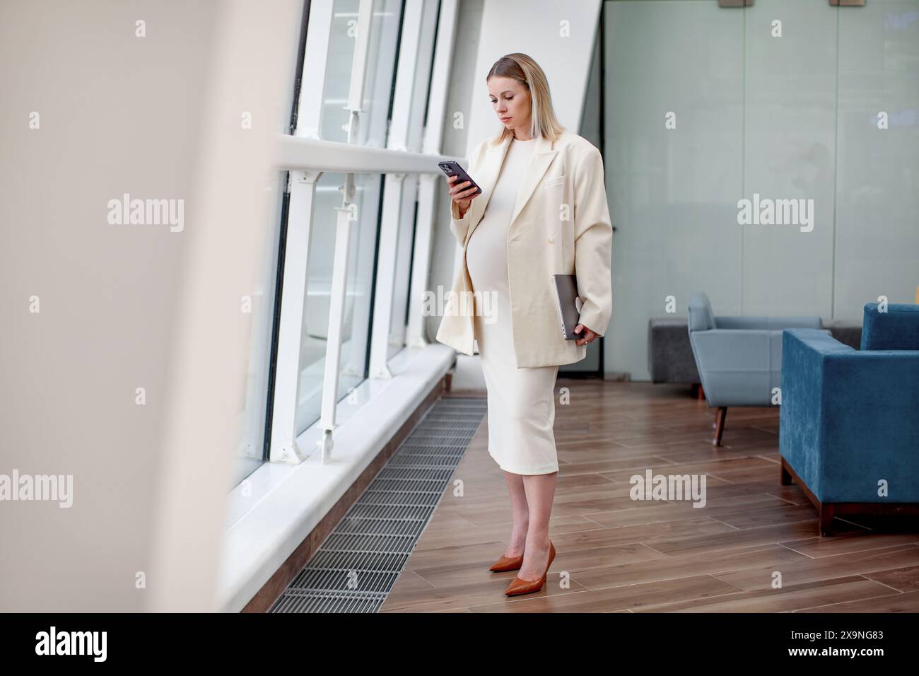Porträt einer schönen schwangeren Geschäftsfrau, die selbstbewusst an einem Laptop arbeitet, in einem modernen Büro mit hellblauem Hintergrund und einem stilvollen Stockfoto