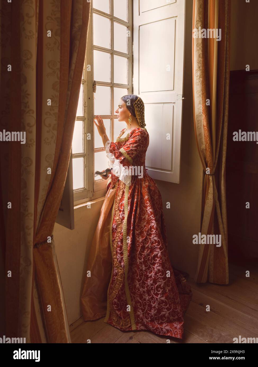 Frau mit authentischem Renaissance-Kostüm und Kopfschmuck, die in einem mittelalterlichen Schloss an ihrem Fenster steht Stockfoto