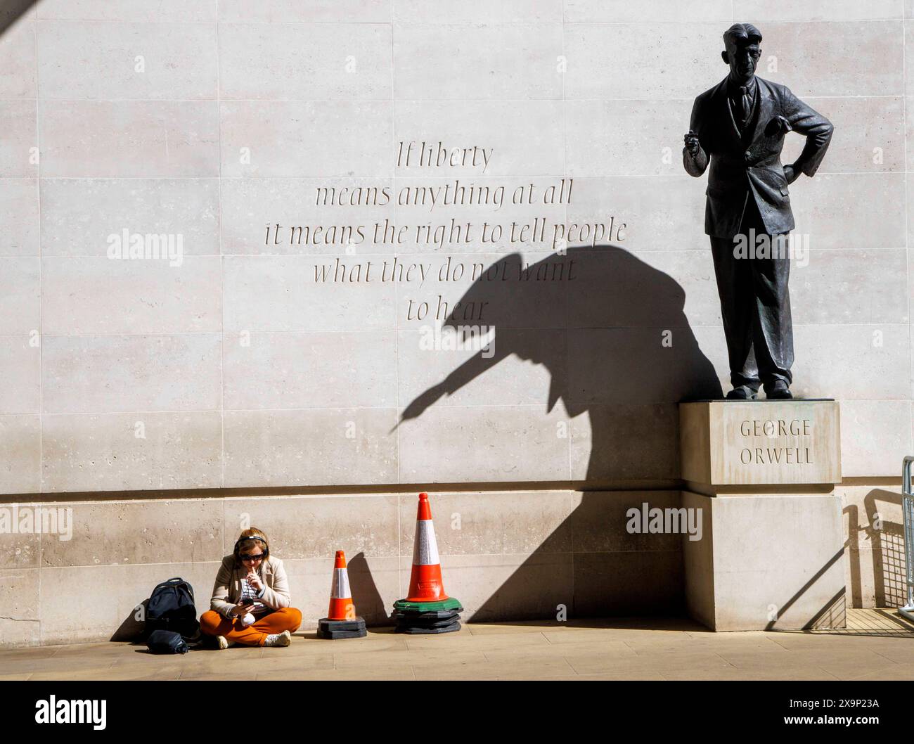 London, Großbritannien. Juni 2024. Eine Frau entspannt sich im Sonnenschein unter einem Schatten der Statue von George Orwell. Die Statue von George Orwell des britischen Bildhauers Martin Jennings wurde am 7. November 2017 vor dem Broadcasting House, dem Hauptquartier der BBC, in London enthüllt. Die Wand hinter der Statue ist mit George Orwells Worten beschriftet. Wenn Freiheit überhaupt etwas bedeutet, bedeutet es das Recht, Menschen zu sagen, was sie nicht hören wollen. Dies war ein Zitat aus einem ungenutzten Vorwort zu Animal Farm. Quelle: Mark Thomas/Alamy Live News Stockfoto