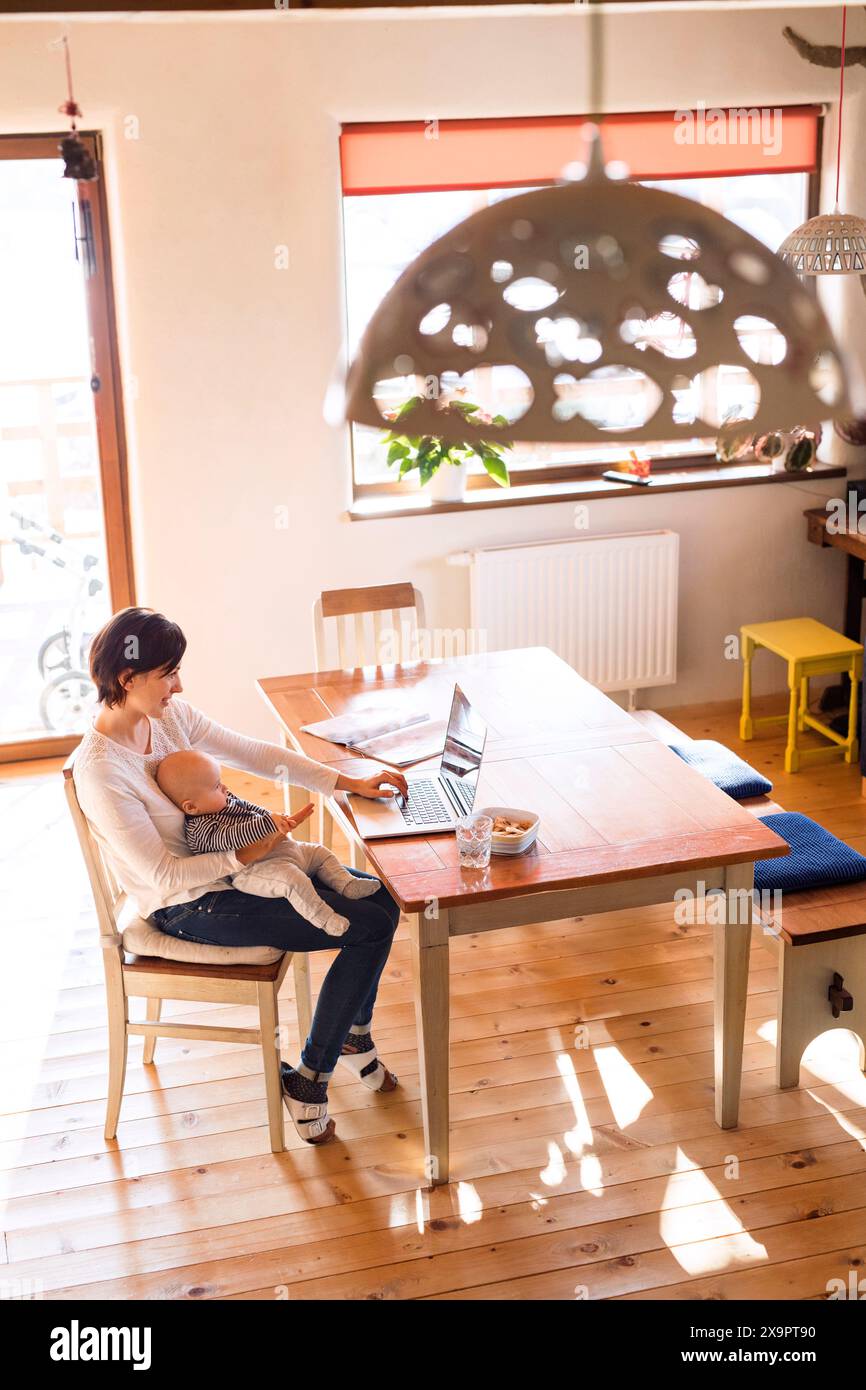 Mutter mit kleinem Baby, die vom Homeoffice aus arbeitet und auf dem Laptop schreibt. Mom bezahlt Rechnungen online, hält ein kleines Baby. Stockfoto