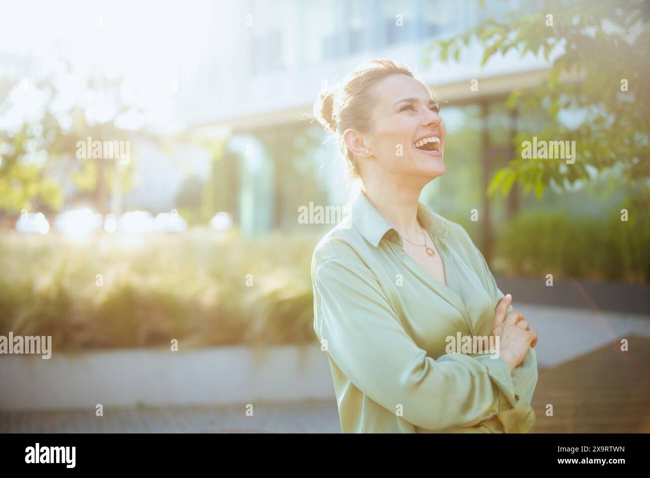 Glückliche moderne Arbeiterin in der Nähe des Bürogebäudes in grüner Bluse mit überkreuzten Armen. Stockfoto