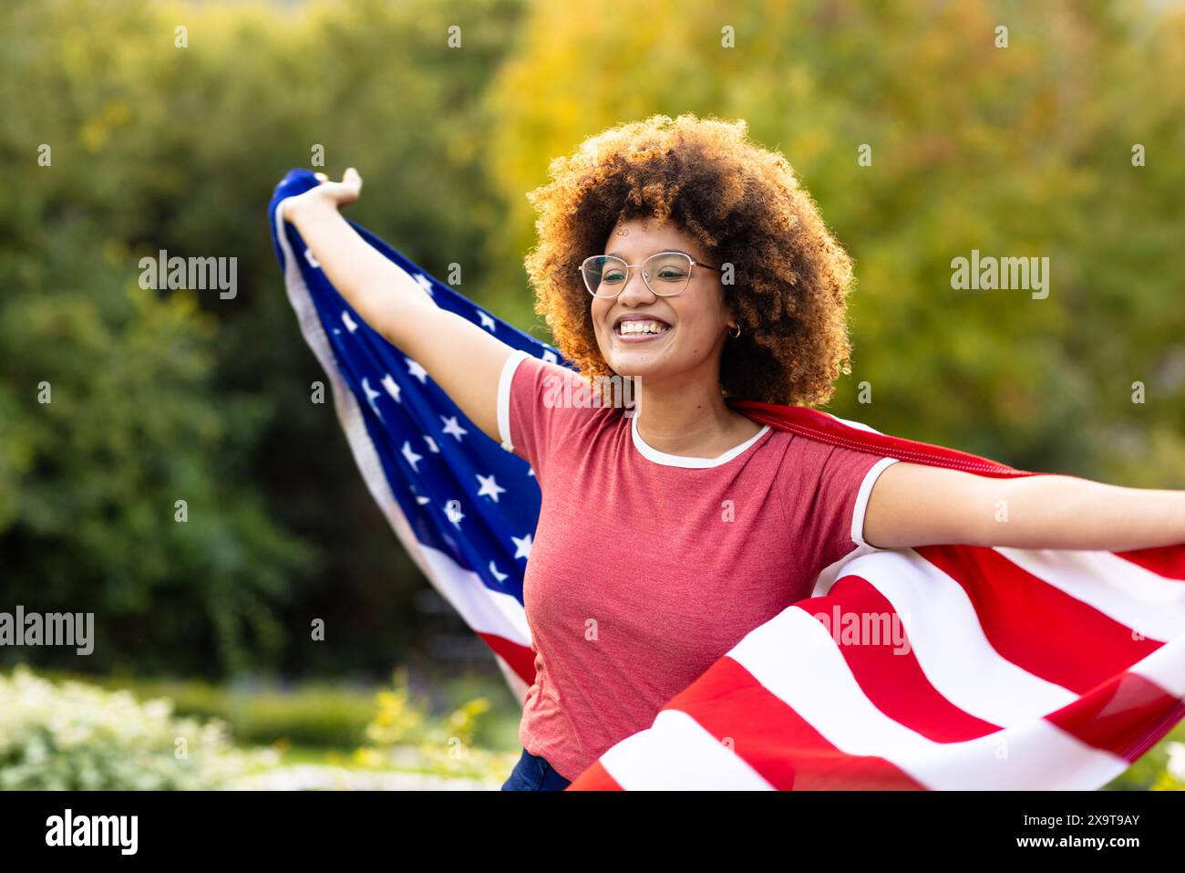 Glückliche Frau, die mit der Flagge der usa bedeckt ist und die Arme weit im Garten liegen Stockfoto