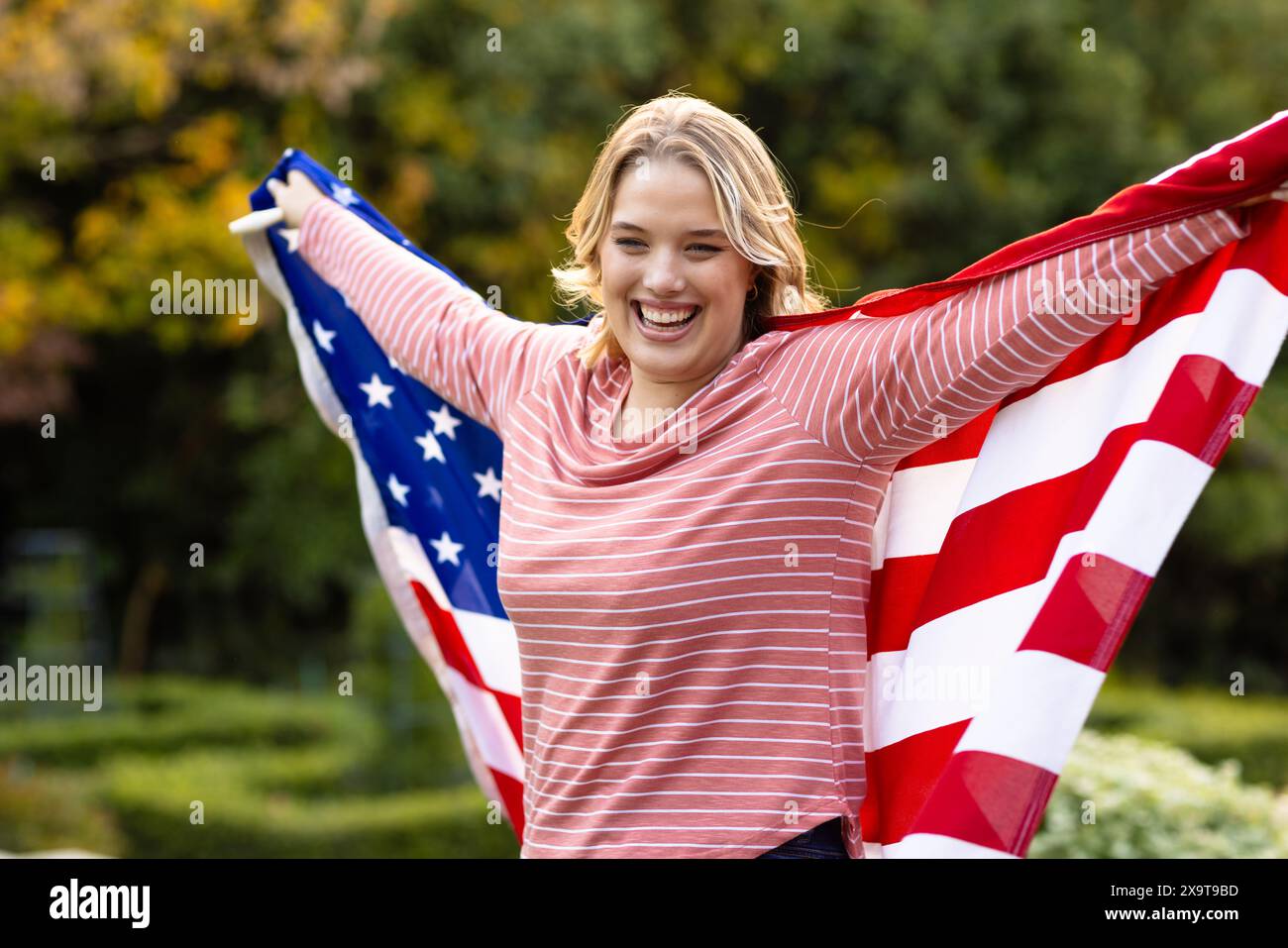Glückliche kaukasische Frau bedeckt mit der Flagge der usa mit weit im Garten liegenden Armen Stockfoto