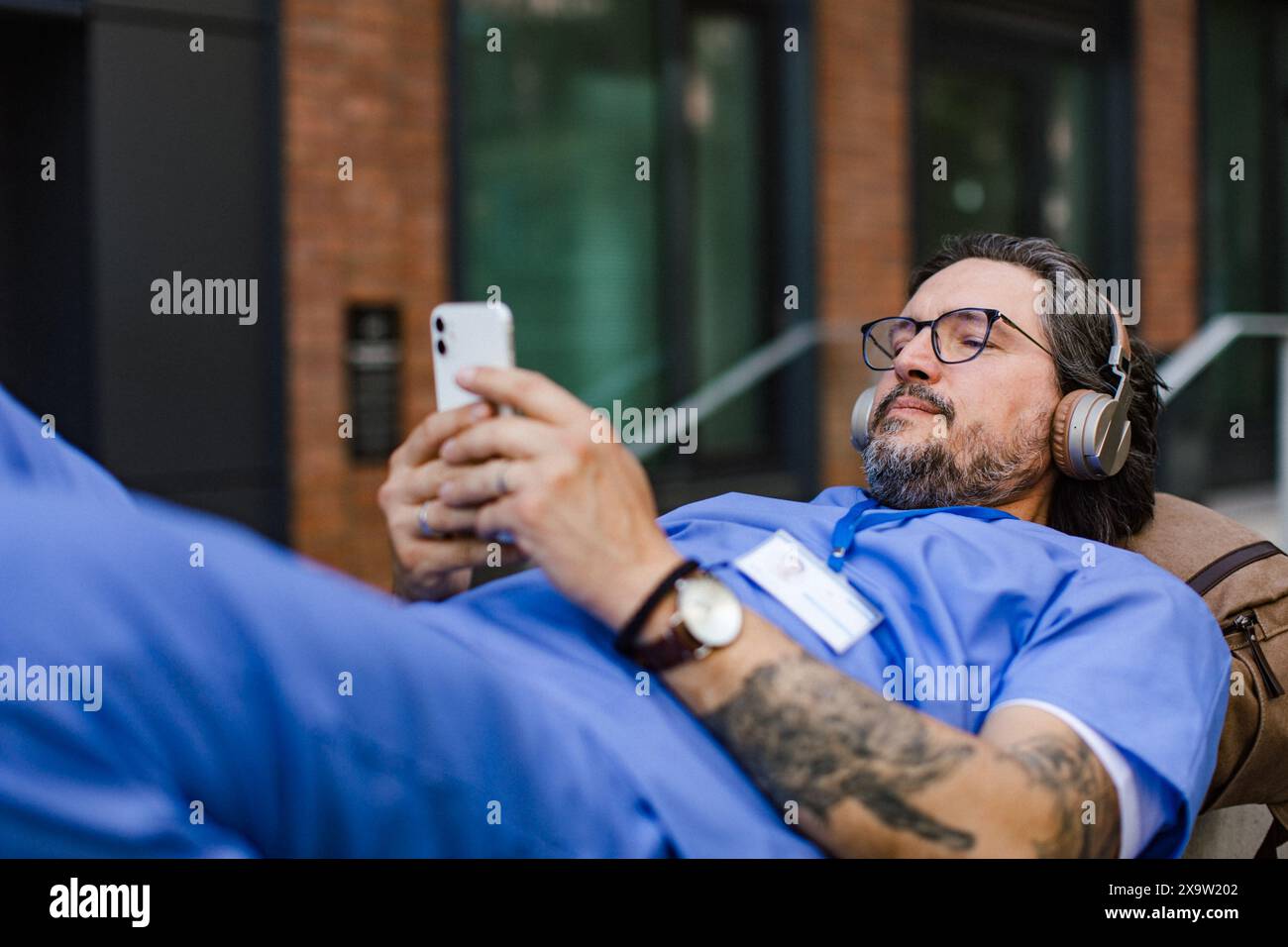 Doktor, der auf der Parkbank liegt, Musik hört, Kopfhörer trägt, auf dem Smartphone scrollt. Nach dem arbeitsreichen Arbeitsalltag im Krankenhaus. Stockfoto