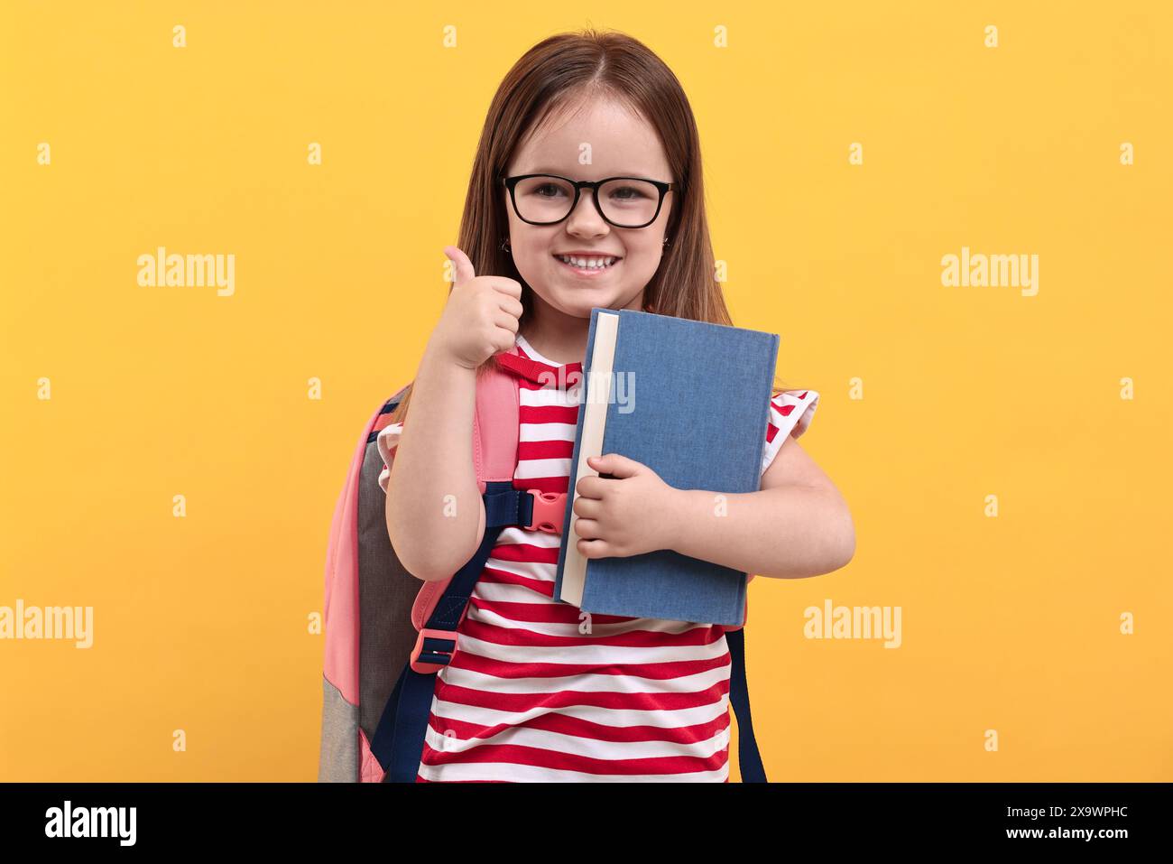 Niedliches kleines Mädchen mit Rucksack und Buch, das die Daumen vor orangem Hintergrund zeigt Stockfoto