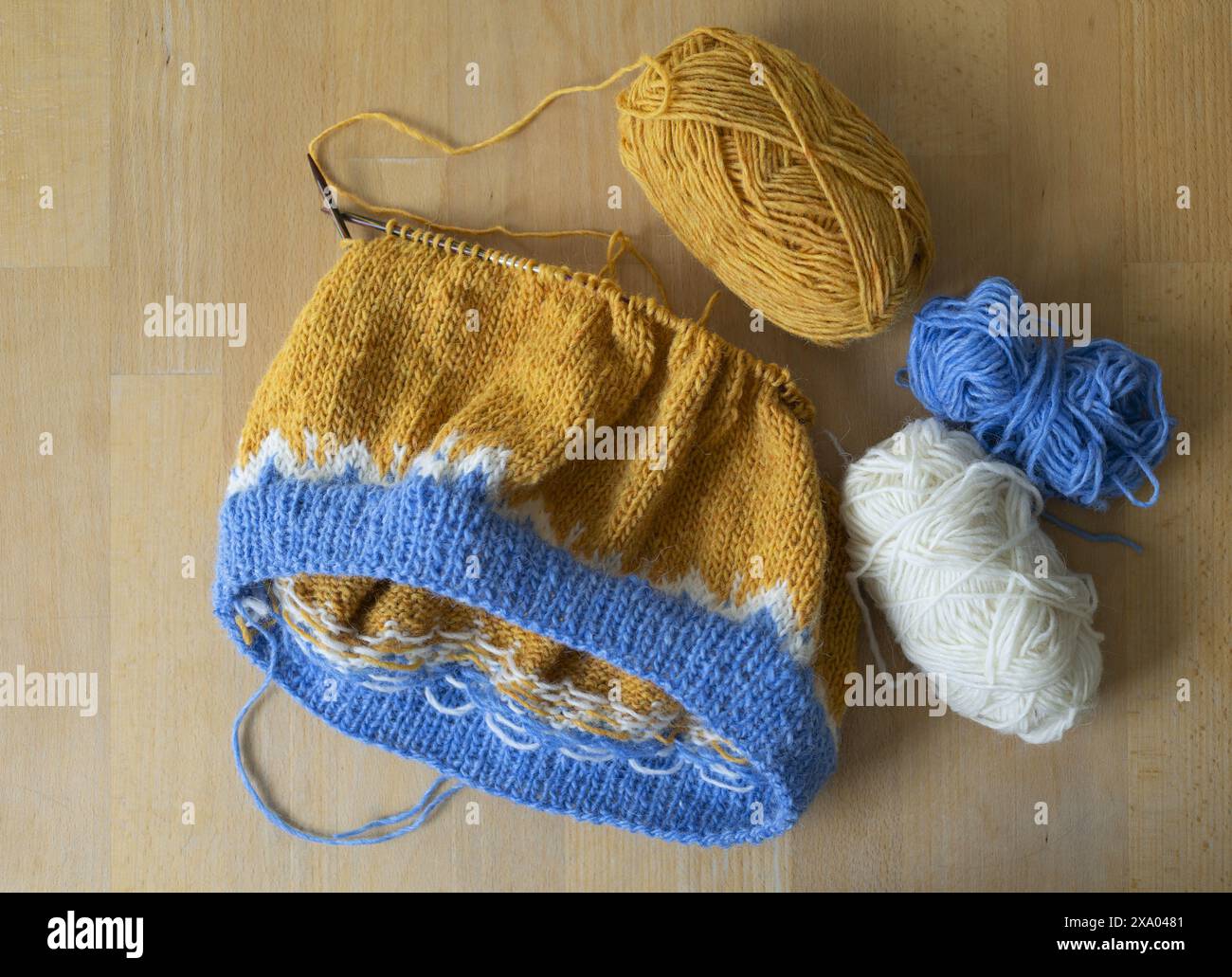 Flachbild-Fotografie eines aus isländischer Wolle gestrickten Lopeysa-Pullovers mit gelben, blauen und weißen Garnbällchen und runden Nadeln auf einem wo Stockfoto