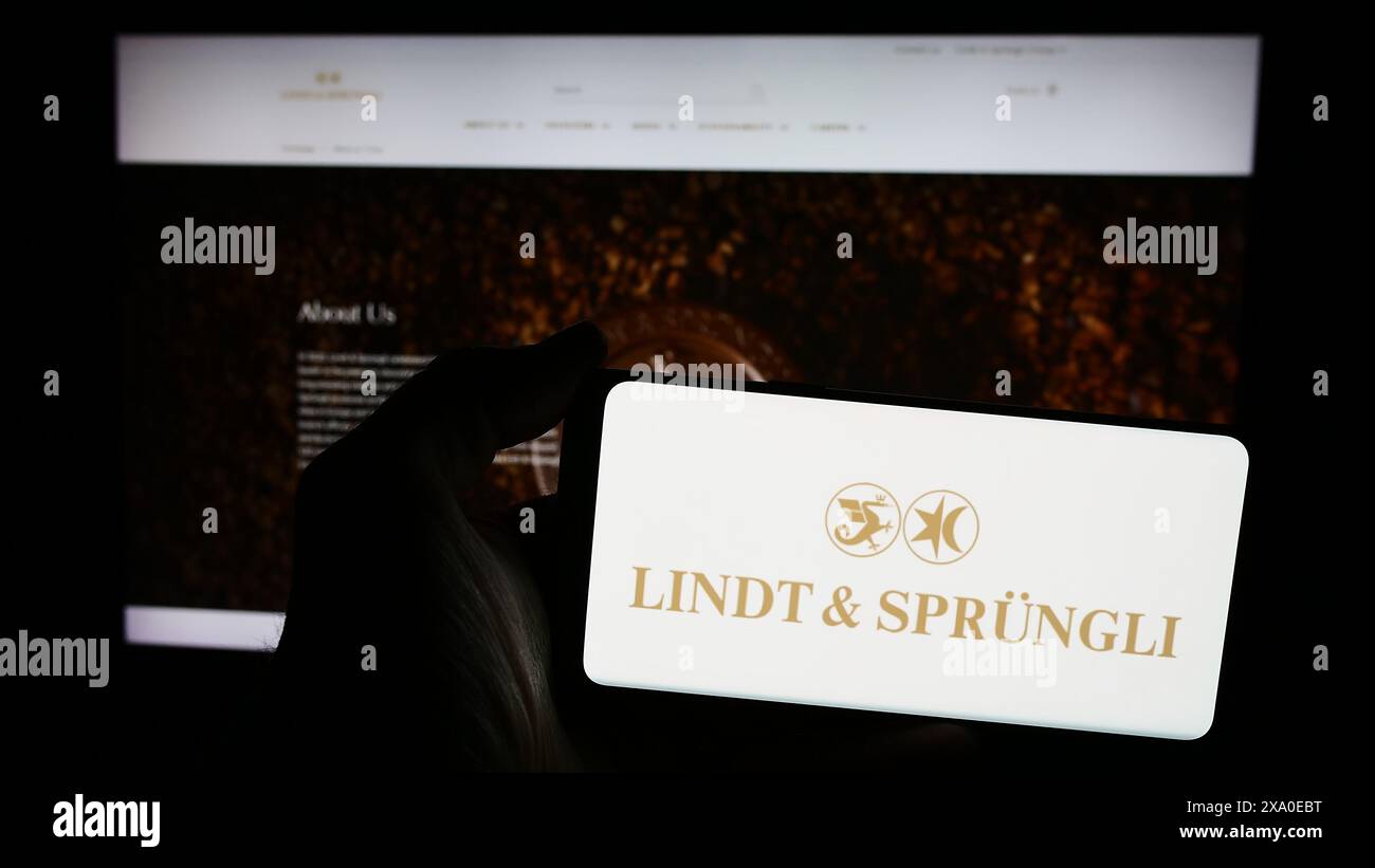 Person mit Smartphone mit Logo der Schweizer Firma Chocoladefabriken Lindt und Sprüngli AG vor der Website. Schwerpunkt auf dem Display des Telefons. Stockfoto