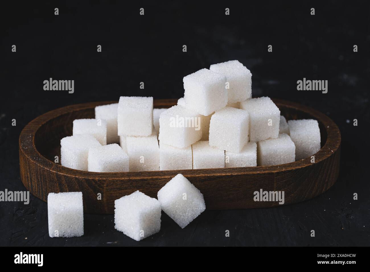 Weiße Würfel aus raffiniertem Zucker auf einem Holzteller. Zuckerwürfel in einer Holzschale auf schwarzem Hintergrund. Stockfoto