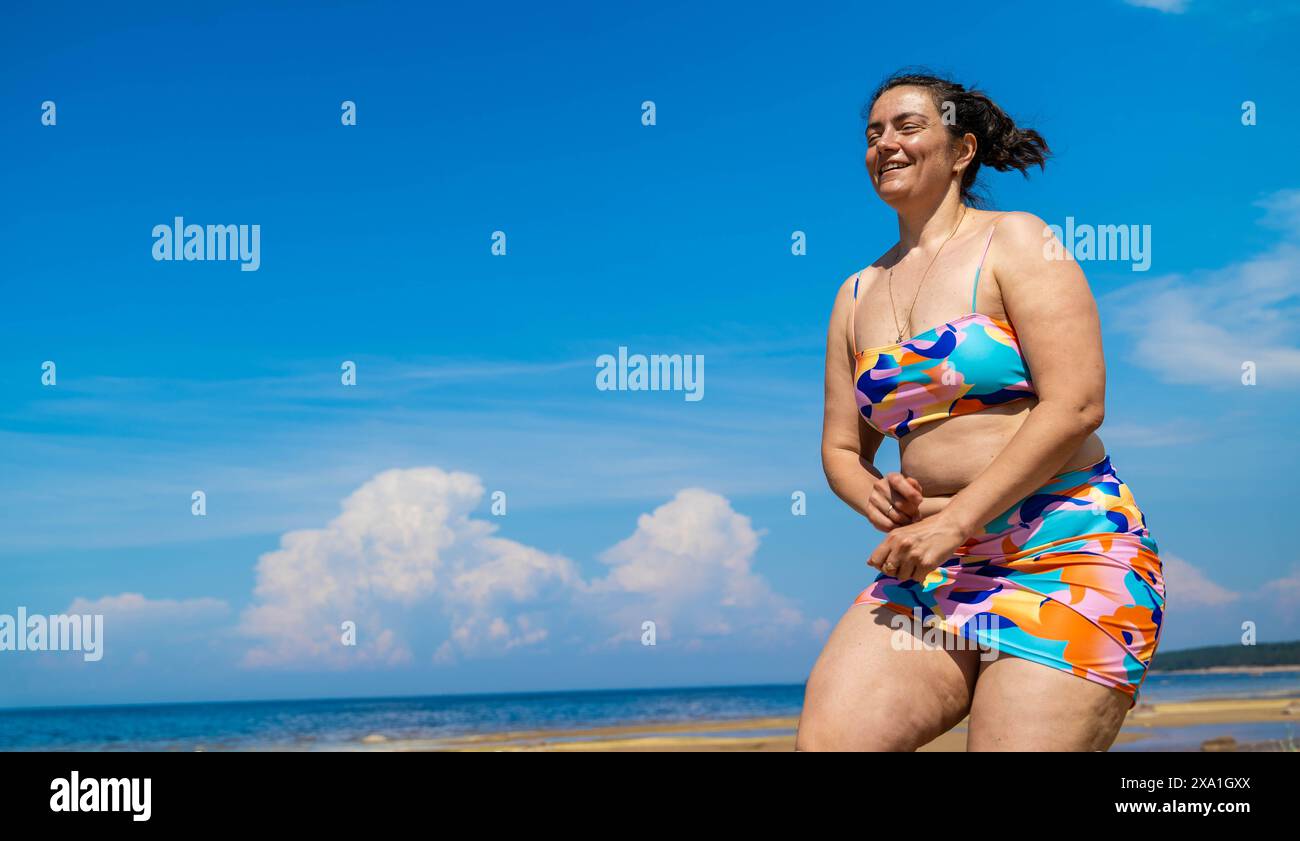 Eine lächelnde Frau im Badeanzug genießt die Sonne am Sandstrand. Körperpositiv, voll, Cellulite Stockfoto Eine lächelnde Frau im Badeanzug genießt die Sonne am Sandstrand. Körperpositiv, voll, Cellulite Stockfoto