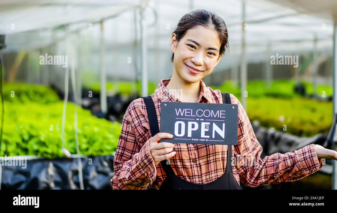 Eine lächelnde asiatische Frau, die in einem Gewächshaus arbeitet und ein Willkommensschild hält Stockfoto