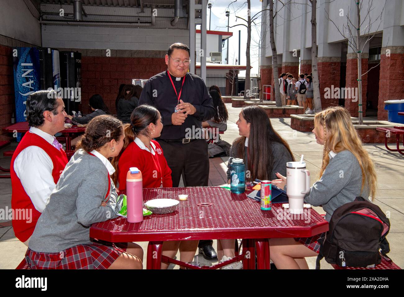 Ein hispanischer Priester unterhält sich mit uniformierten Schülern an einer katholischen Highschool in Südkalifornien, während sie ihr Mittagessen im Freien essen. Stockfoto