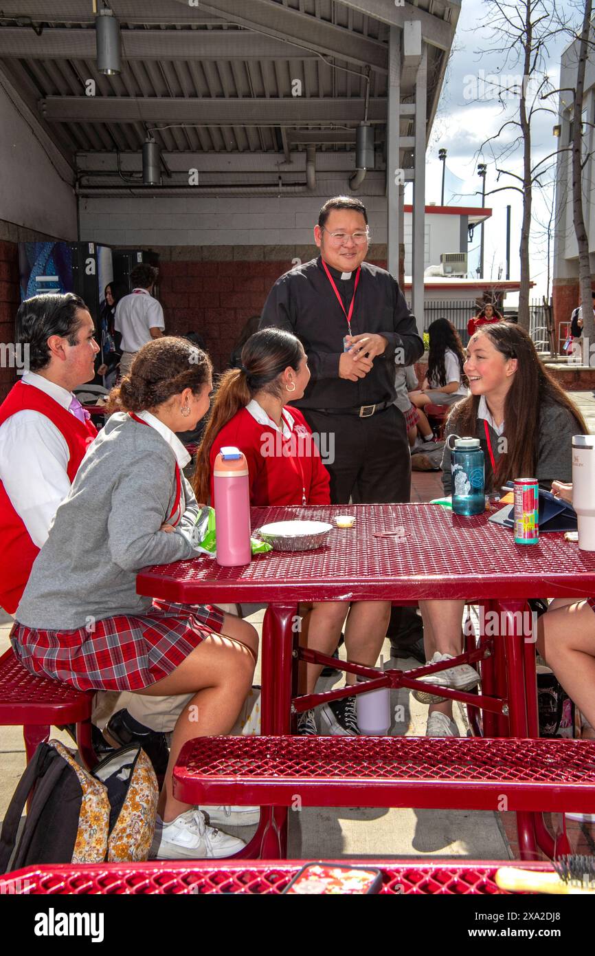 Ein hispanischer Priester unterhält sich mit uniformierten Schülern an einer katholischen Highschool in Südkalifornien, während sie ihr Mittagessen im Freien essen. Stockfoto