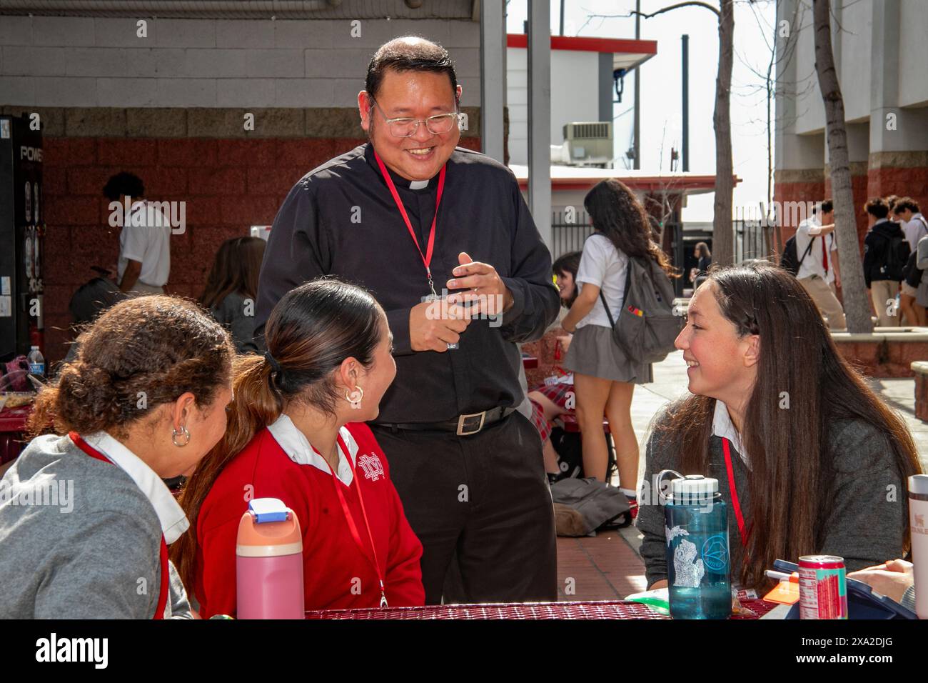 Ein hispanischer Priester unterhält sich mit uniformierten Schülern an einer katholischen Highschool in Südkalifornien, während sie ihr Mittagessen im Freien essen. Stockfoto