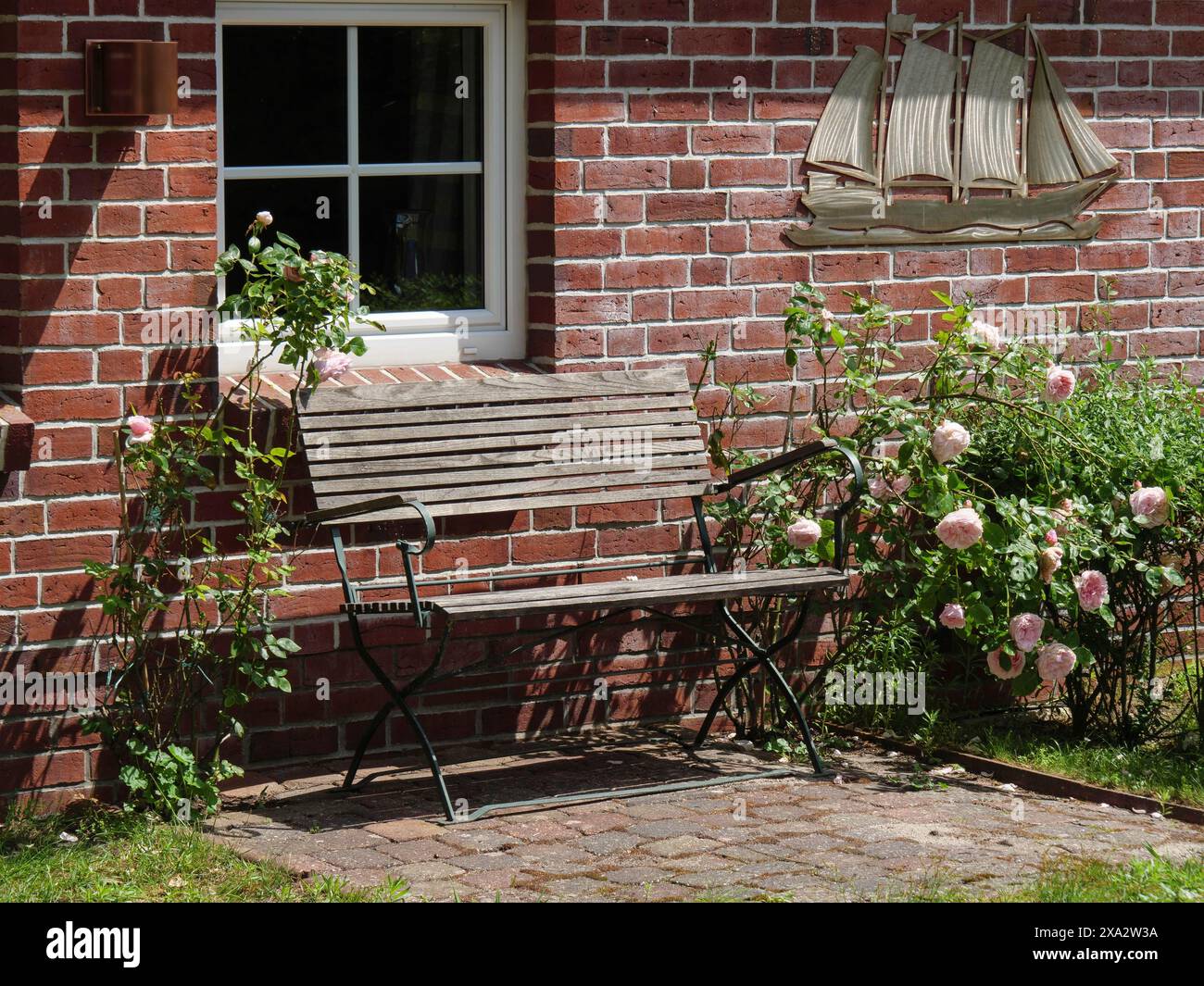 Eine Holzbank neben Rosenbüschen vor einer roten Ziegelmauer mit Fenster, Baltrum Deutschland Stockfoto