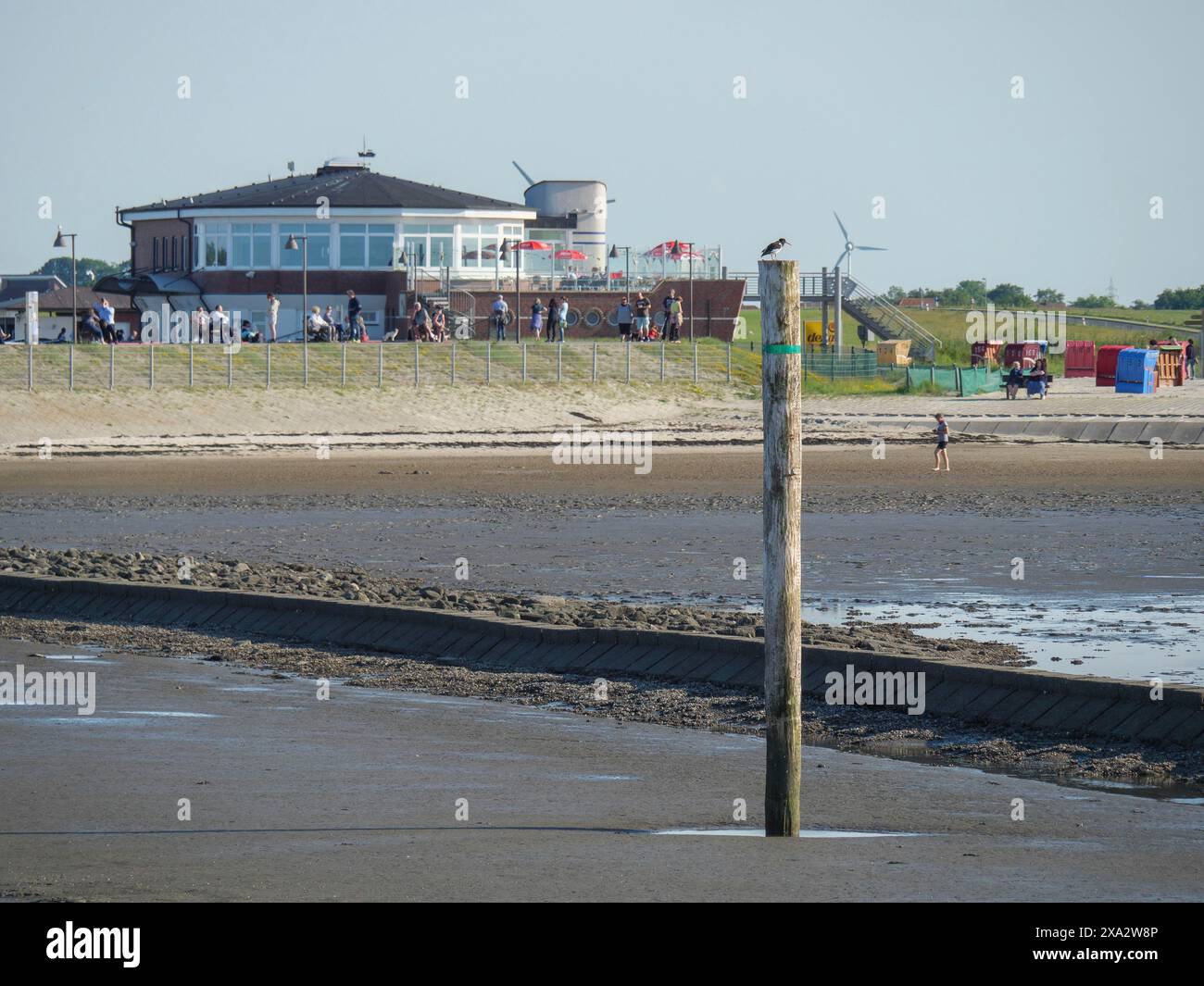 Eine Stange am Strand mit Wasser davor, im Hintergrund ein modernes Gebäude und Liegen, Menschen am Strand, Baltrum Deutschland Stockfoto