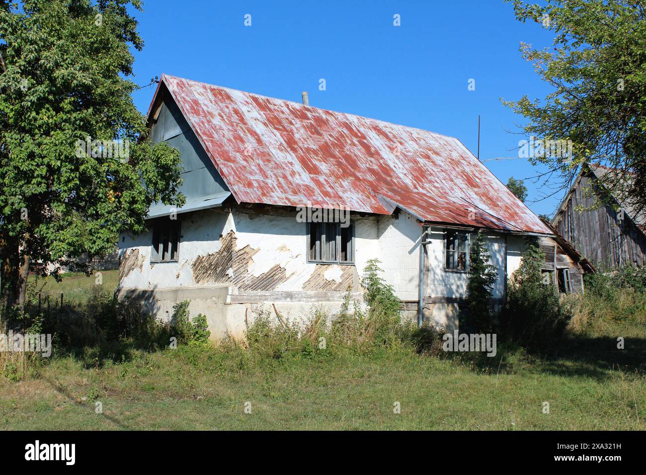 Verlassenes kleines altes urbanes Familienhaus mit zerstörter weißer Fassade um baufällige Holzrahmenfenster mit Glasbruch, die mit Rot bedeckt sind Stockfoto