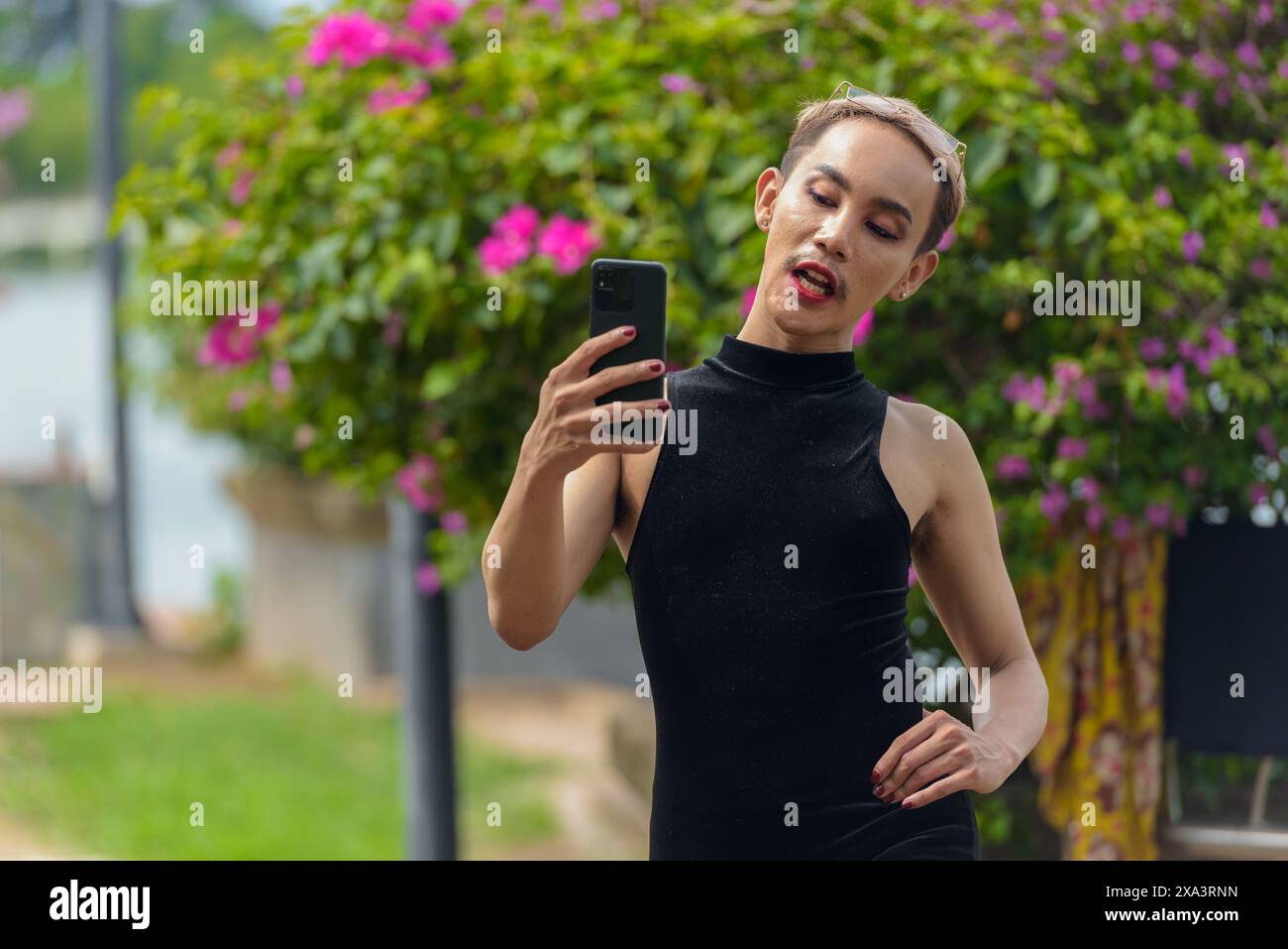 Wunderschöner asiatischer queerer LGBT-Community-Unterstützer Mann mit Schnurrbart, der Lippenstift und Jumpsuit trägt und draußen im Park telefoniert Stockfoto