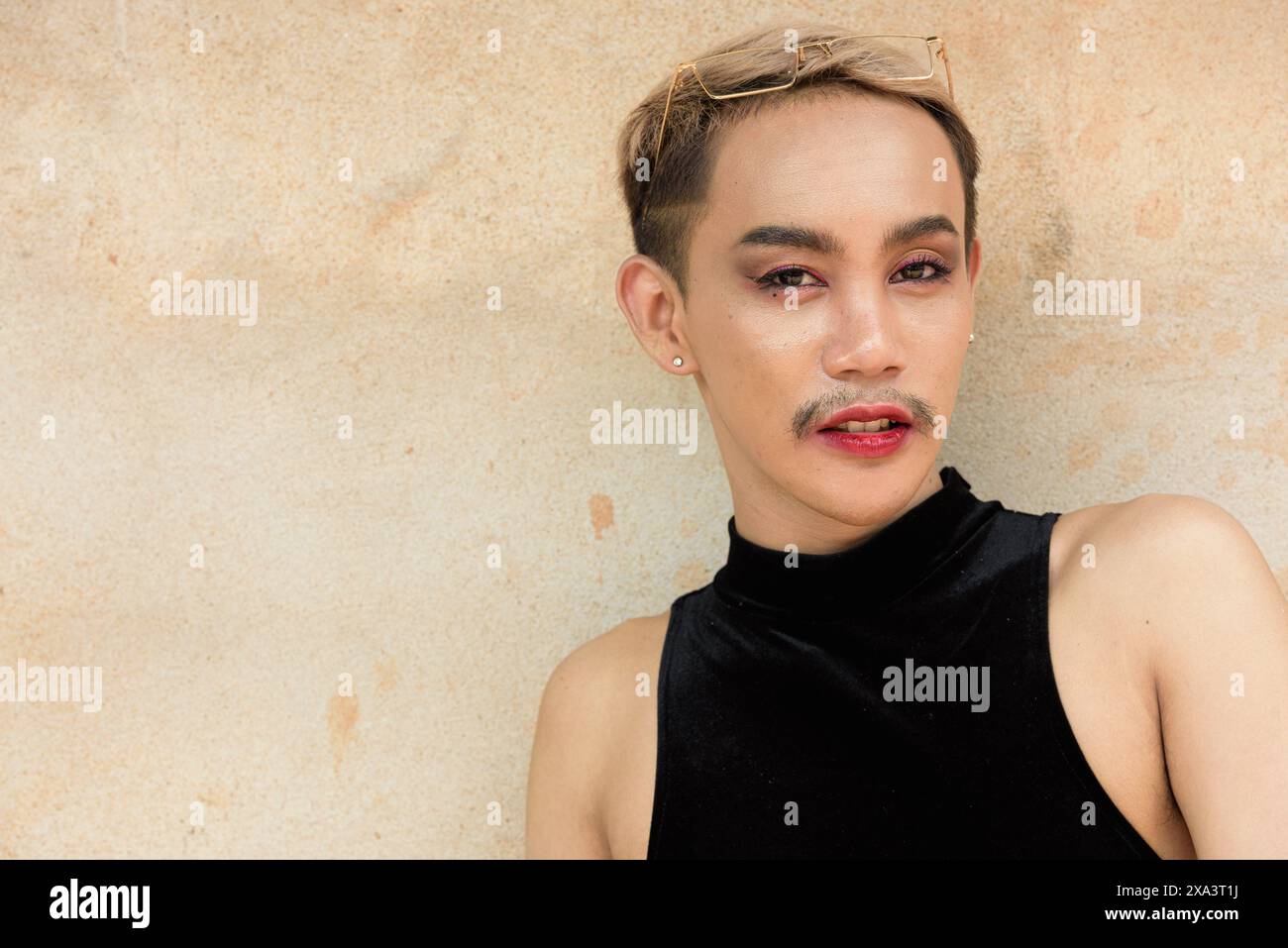 Porträt des schönen asiatischen queeren LGBT Community Unterstützers Mann mit Schnurrbart, der Lippenstift und Jumpsuit vor beigem Wandhintergrund trägt Stockfoto