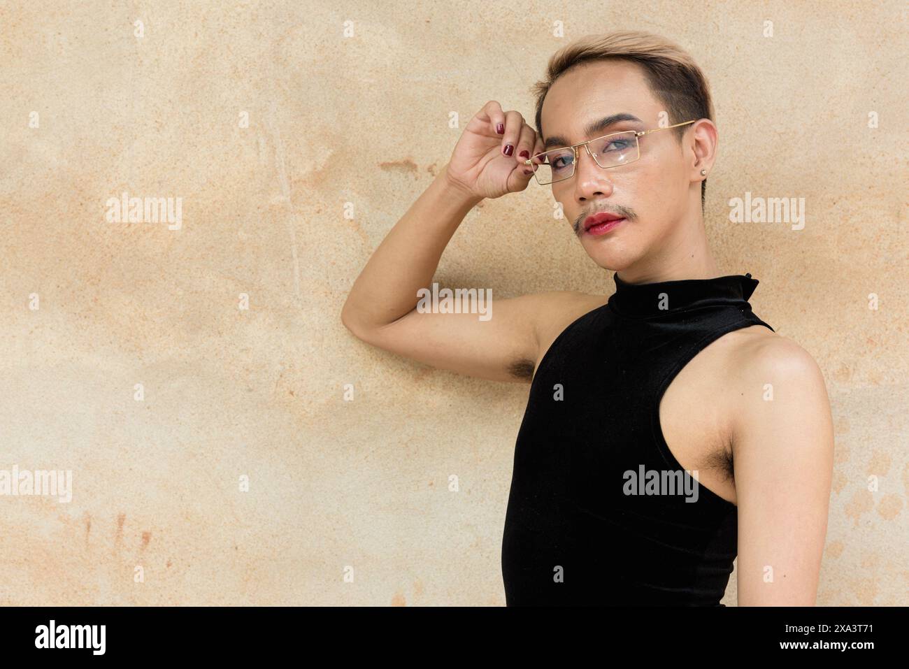 Porträt des schönen asiatischen queeren LGBT Community Unterstützers Mann mit Schnurrbart, der Lippenstift und Jumpsuit vor beigem Wandhintergrund trägt Stockfoto