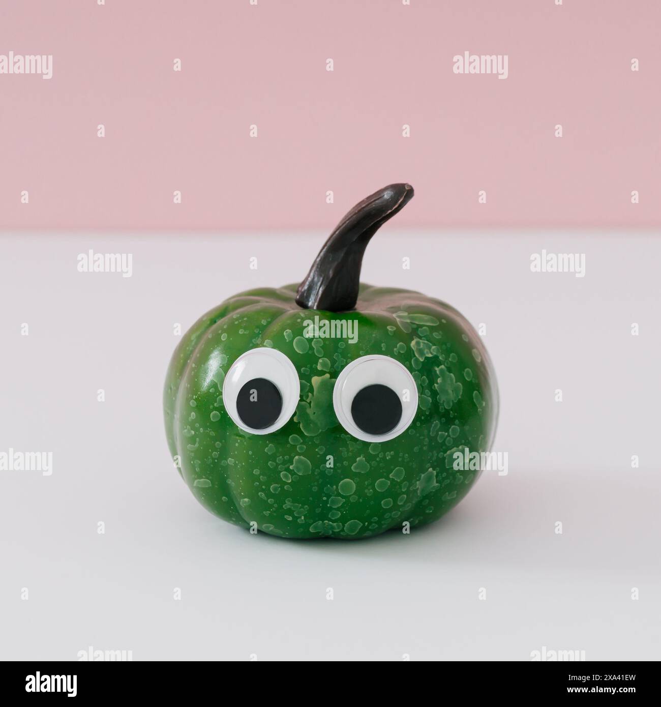 Süßer kleiner grüner Kürbis mit großen Augen auf weißem und rosa Hintergrund. Minimaler Hintergrund für Herbst- oder Herbstsaison. Lustige Kürbiskomposition. Stockfoto