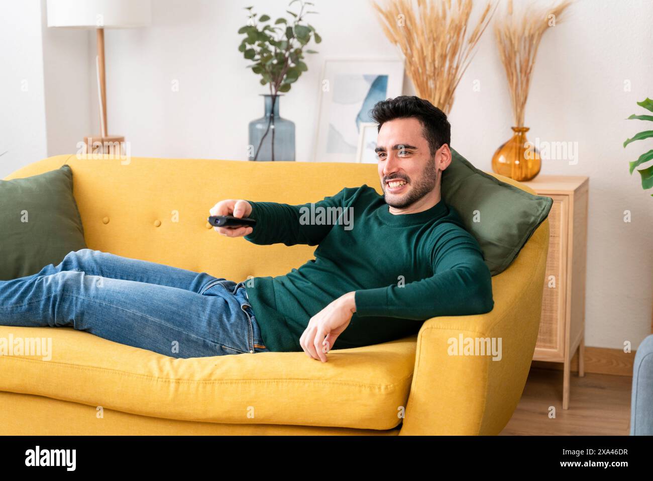 Mann, der sich auf einem gelben Sofa mit Fernbedienung entspannt Stockfoto