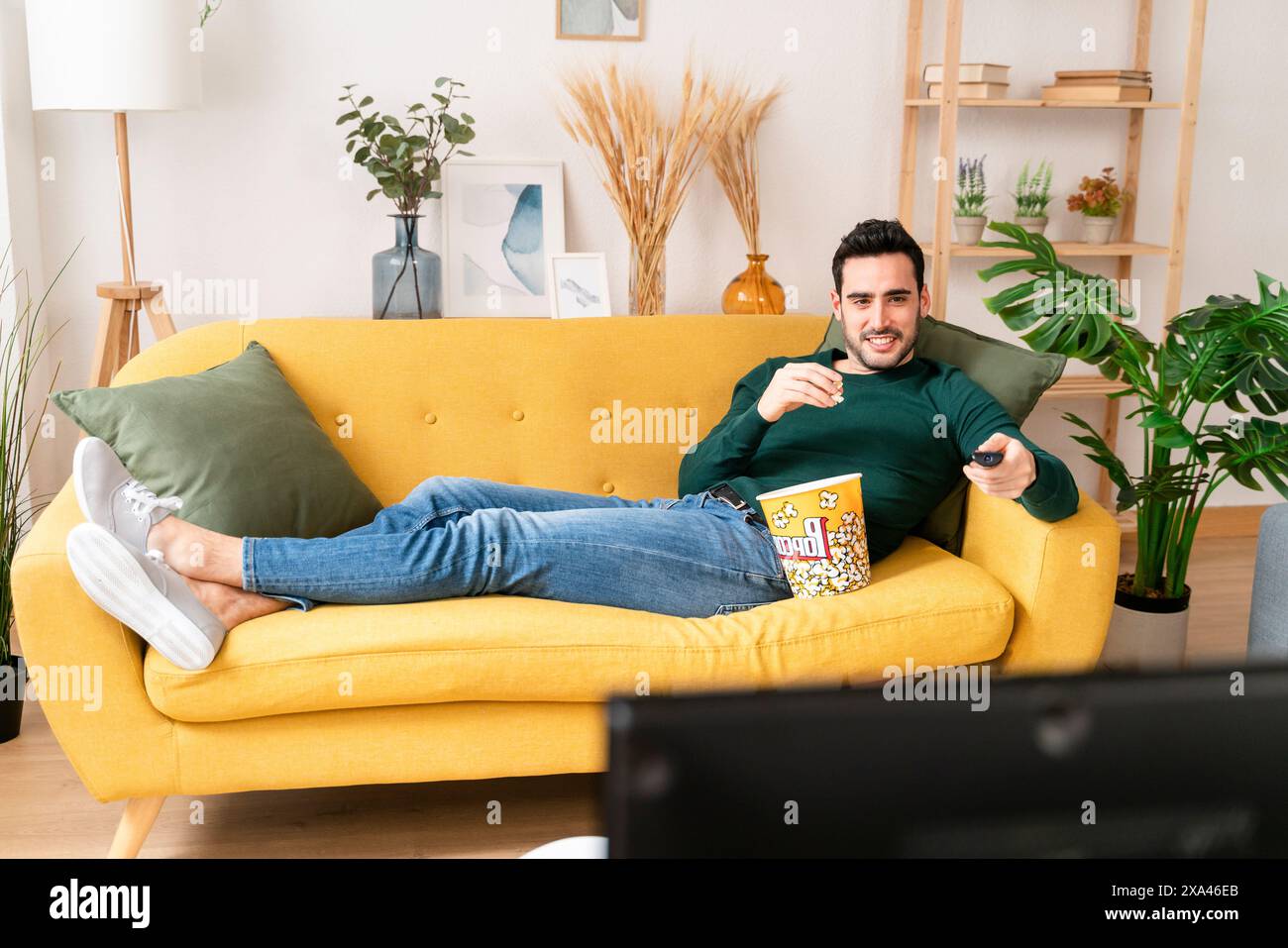 Mann, der sich auf einem gelben Sofa mit einem Snack und Fernbedienung entspannt Stockfoto