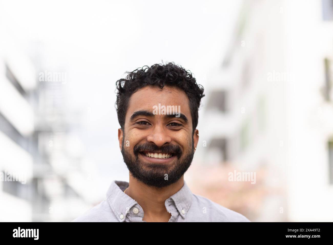 Ein fröhlicher Mann mit Bart und lockigem Haar lächelt vor einem verschwommenen urbanen Hintergrund in die Kamera. Stockfoto