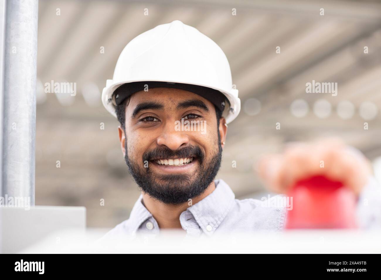 Ein lächelnder Mann in einem weißen Schutzhelm drückt einen roten Knopf, mit einer Stahlkonstruktion im Hintergrund. Stockfoto