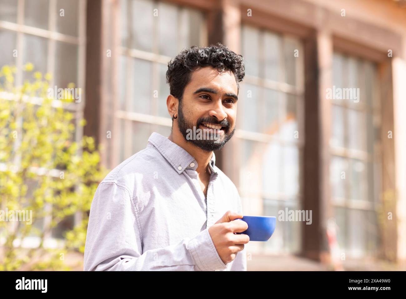 Ein lächelnder Mann mit Bart hält draußen eine blaue Tasse, steht in einem sonnigen Bereich Stockfoto
