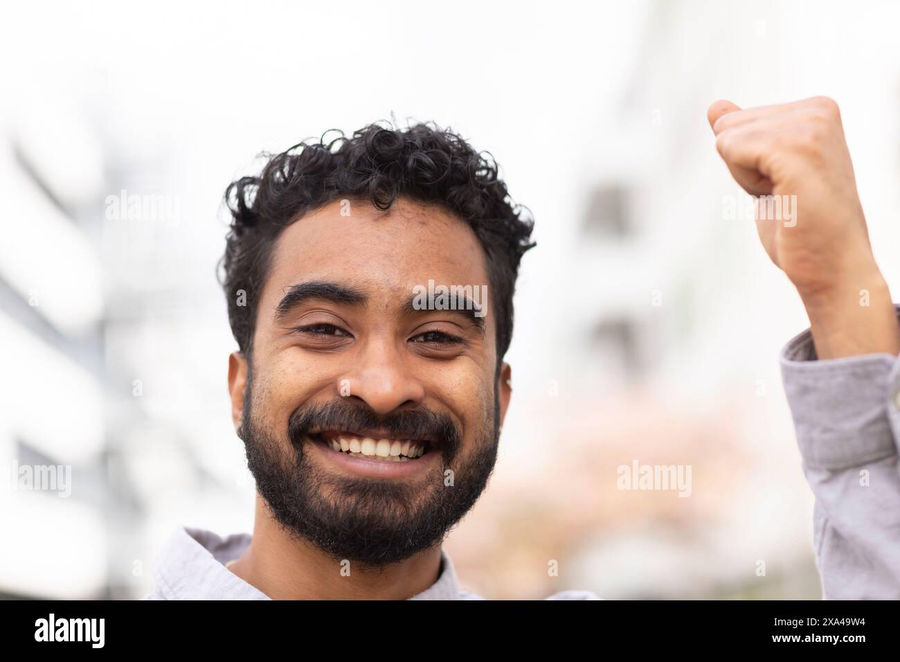 Ein lächelnder Mann mit lockigen Haaren hebt seine Faust in einer Geste des Erfolgs oder der Feier vor einem verschwommenen städtischen Hintergrund. Stockfoto