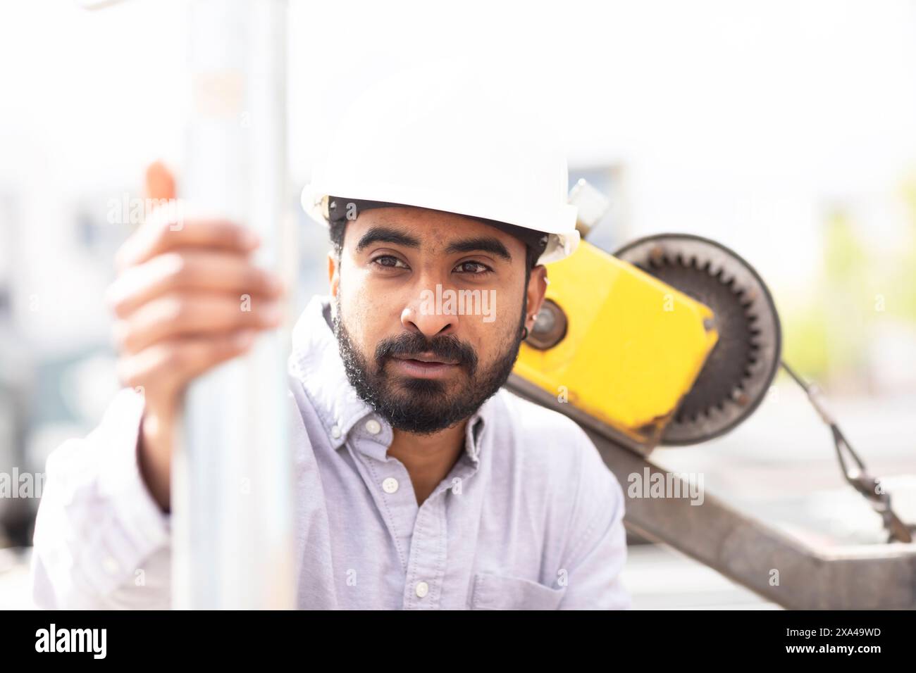 Ein Bauarbeiter in weißem Schutzhelm und Hemd greift einen Metallmast mit gelben Baumaschinen im Hintergrund. Stockfoto